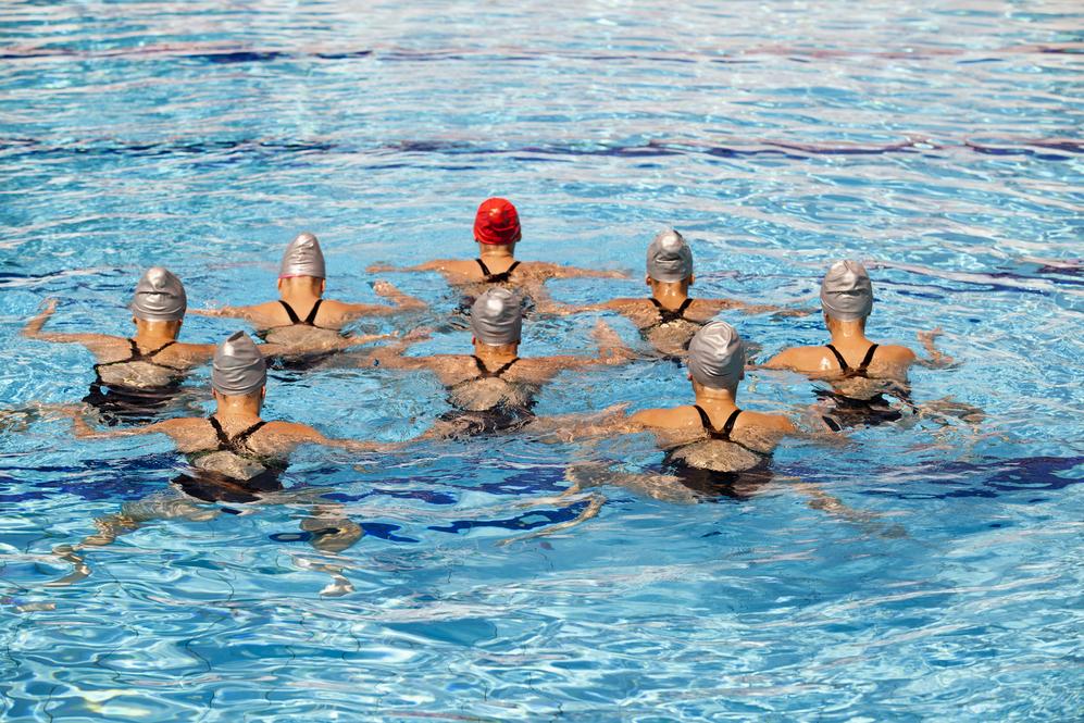 Gruppe von Synchron-Schwimmerinnen in Badeanzügen und Badekappen, die in einem Schwimmbecken synchron schwimmen.
