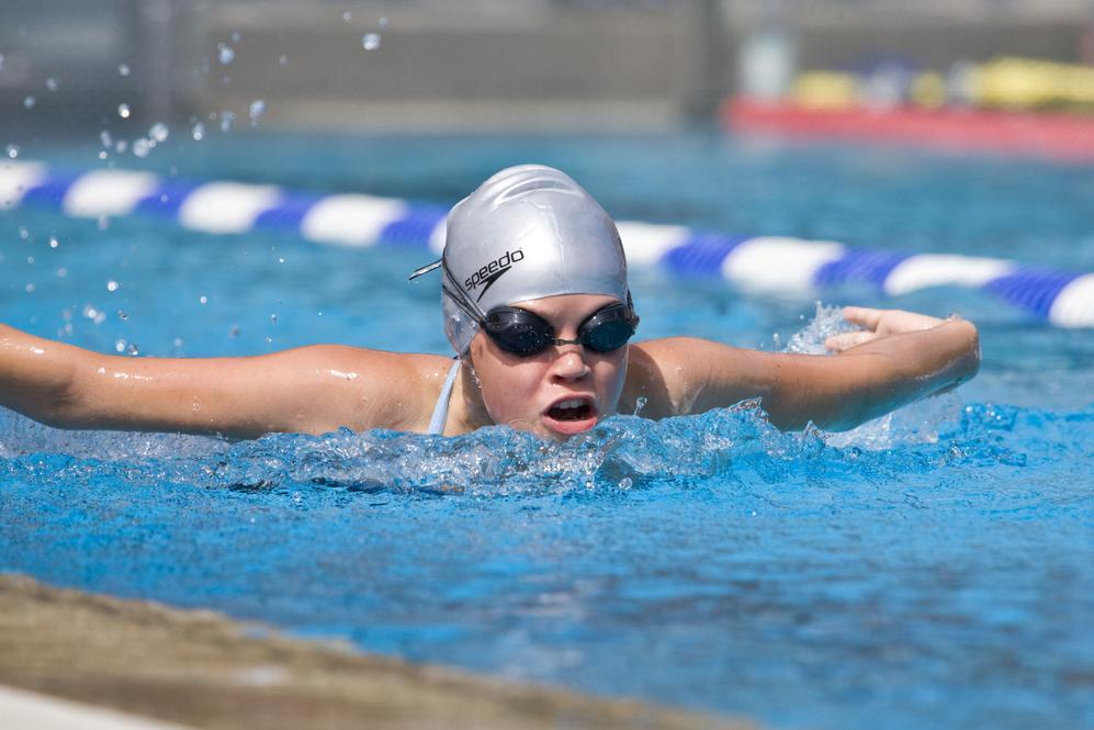Junge Schwimmerin mit silberner Badekappe und Schwimmbrille in einem Wettkampfbecken, während sie einen Schlag ausführt.