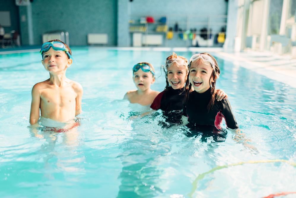 Vier Kinder mit Schwimmbrillen stehen lachend im Wasser eines Schwimmbads. Zwei tragen Neoprenanzüge.