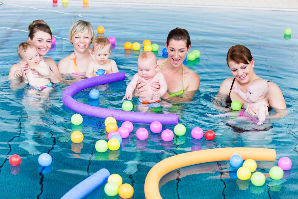 Vier Frauen stehen im Wasser und halten Babys, umgeben von bunten Bällen und Schaumstoffringen in einem Schwimmbad.